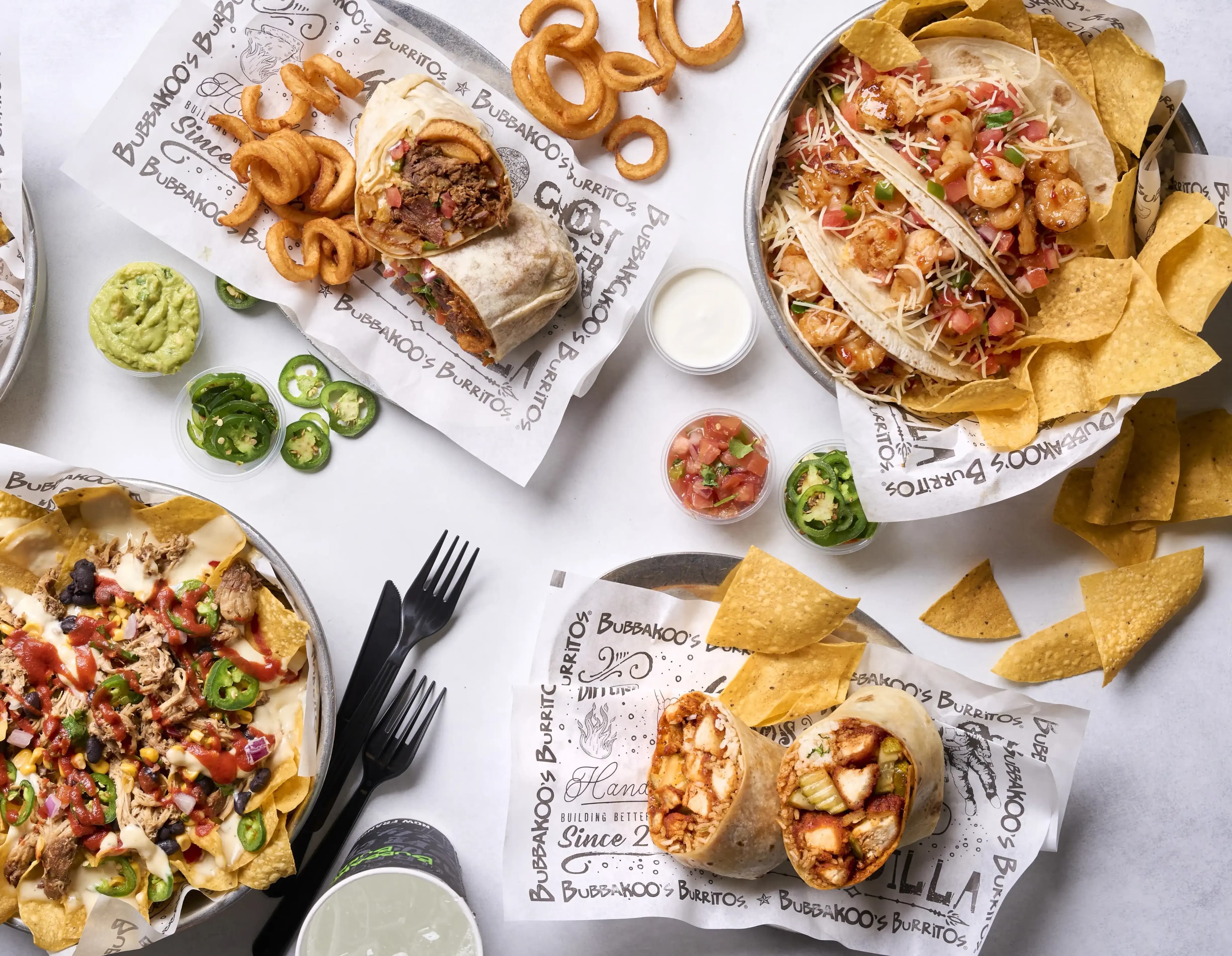 Assorted Mexican Fusion dishes from Bubbakoo's Burritos, including tacos, burritos, nachos with cheese and jalapeños, curly fries, tortilla chips, guacamole, and dipping sauces.