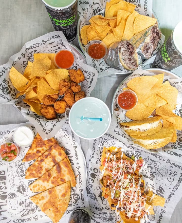 Assorted Bubbakoo’s Burritos catering spread including nachos, quesadillas, tacos, burritos, chips, sauces, and fountain drinks served on branded paper.